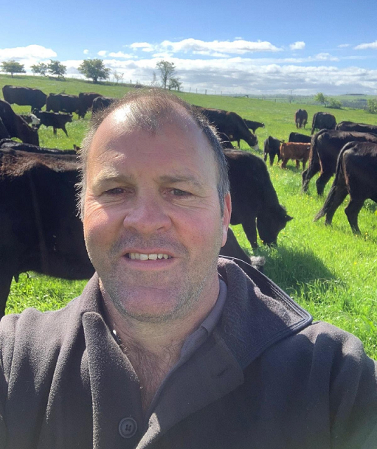 A man (Angus Nelless) stood in a grass field with dark coloured cows behind him